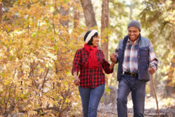 A middle-aged couple are walking in the forest in the fall, surrounded by autumn colors. The man is using a wooden stick for balance.