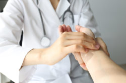 A female doctor in a white coat with a stethoscope around her neck massages the hand of a patient.